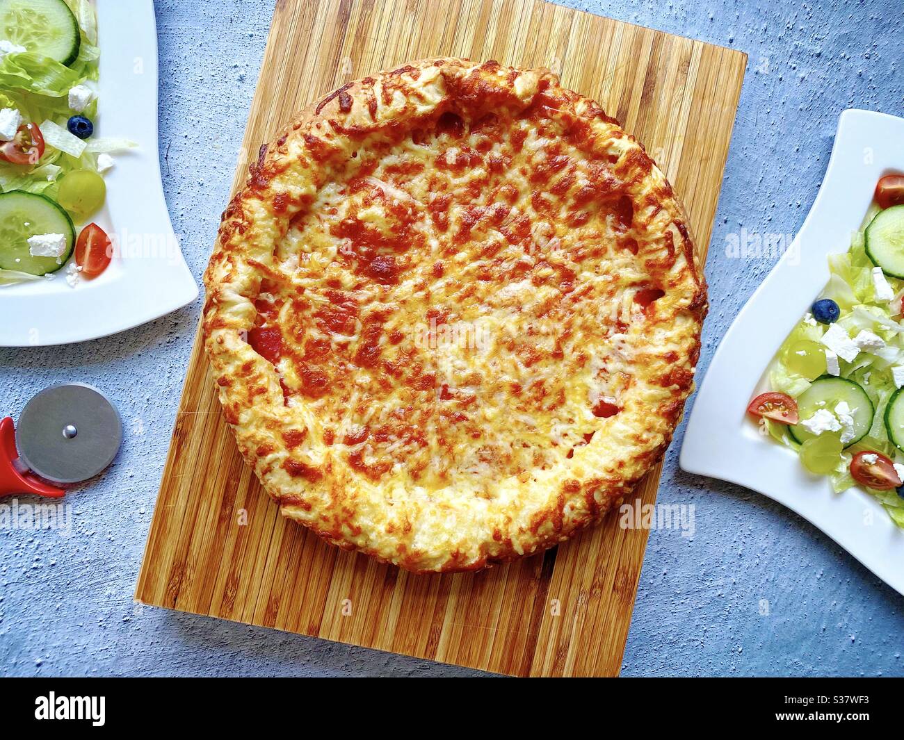 Top down view of freshly baked deep pan cheese and tomato pizza with two plates of fresh feta salad. Cheesy pizza with thick crust against a rough concrete kitchen counter. Flat lay food photography. Stock Photo