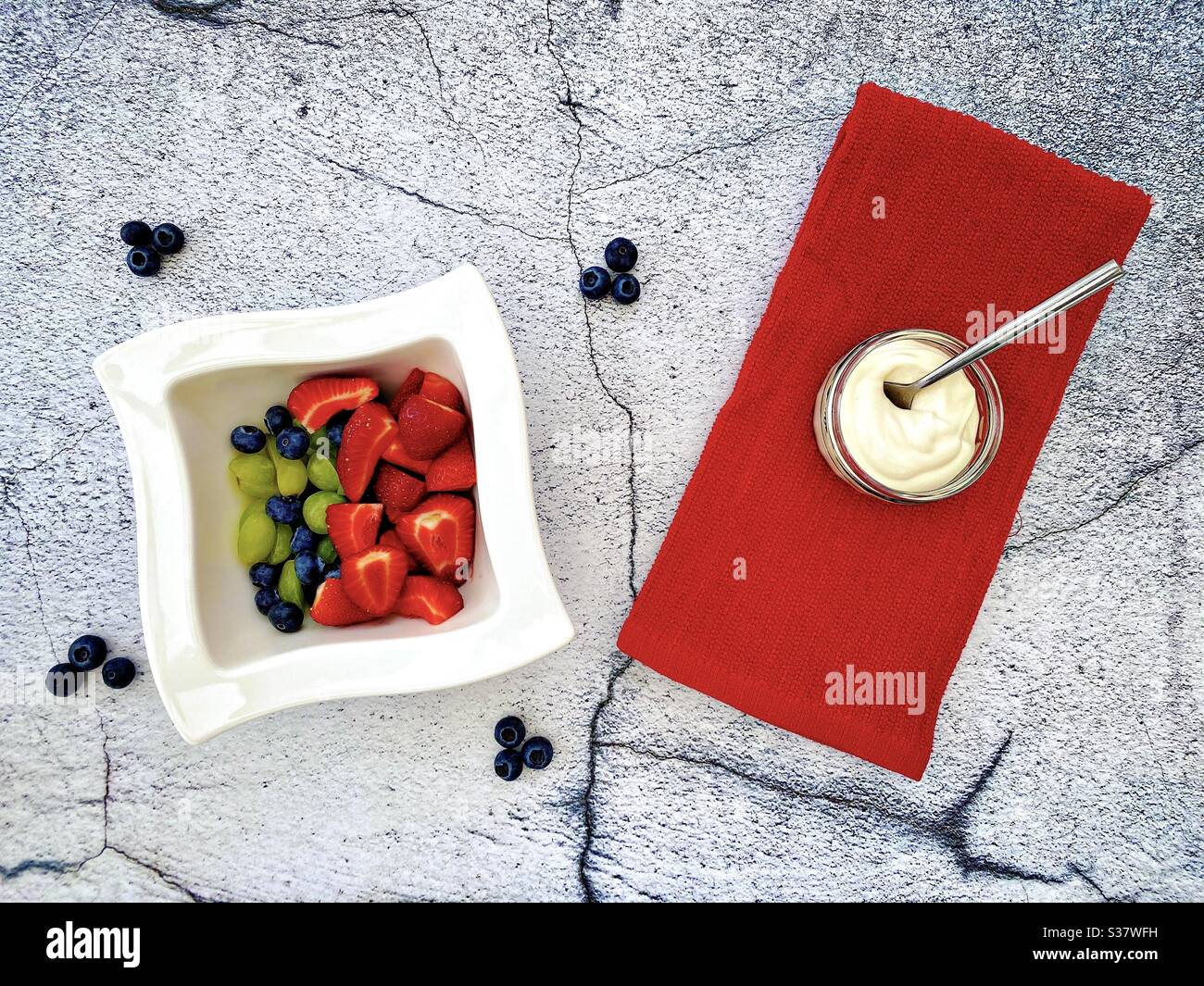 Closeup view of fresh summer berries in a white bowl against a textured natural cracked stone background in the kitchen. Preparing strawberries, blueberries and grapes with Greek yogurt. - Smartphone Captured Stock Image
