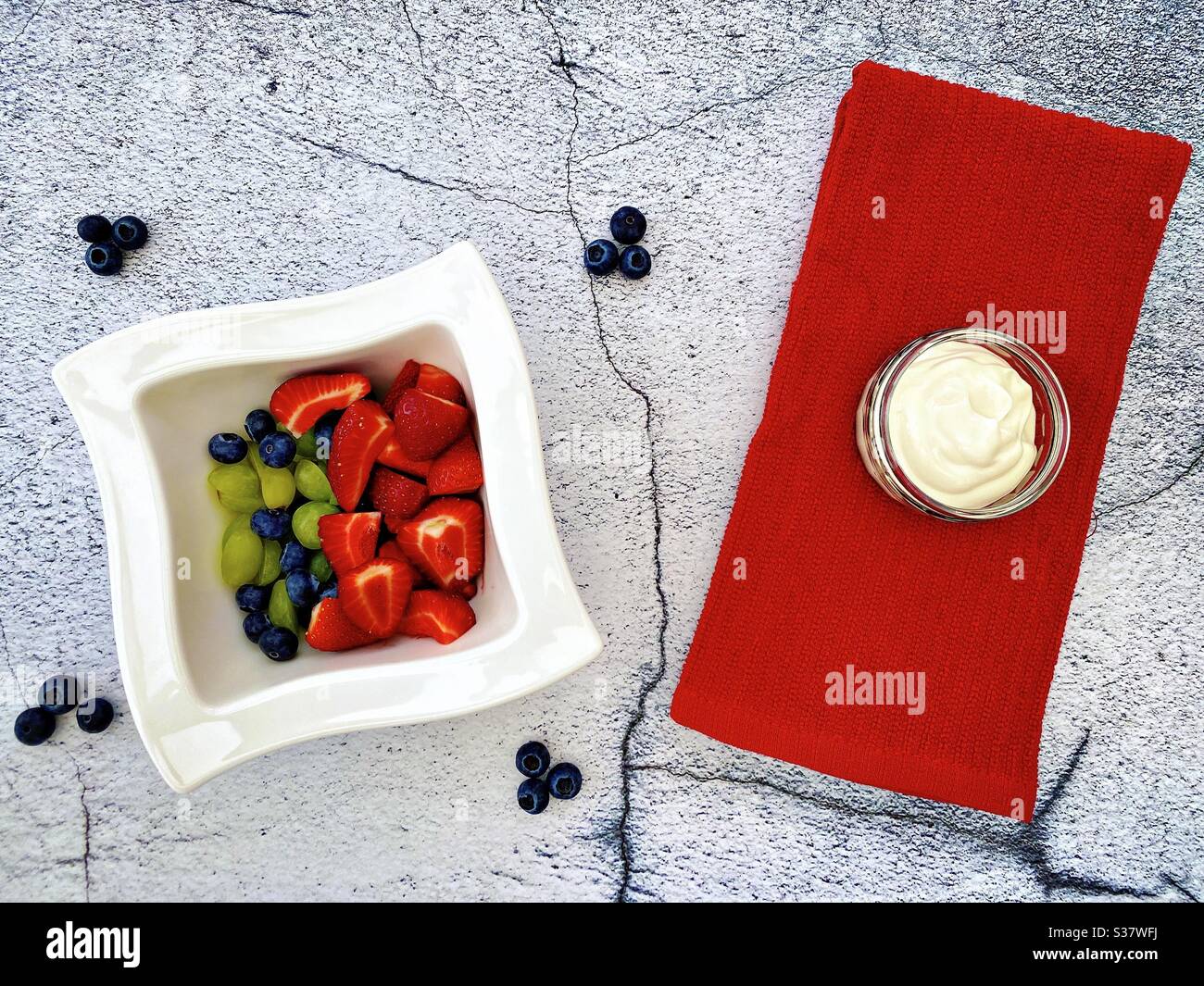 Closeup view of fresh summer berries in a white bowl against a textured natural cracked stone background in the kitchen. Preparing strawberries, blueberries and grapes with Greek yogurt. - Smartphone Captured Stock Image