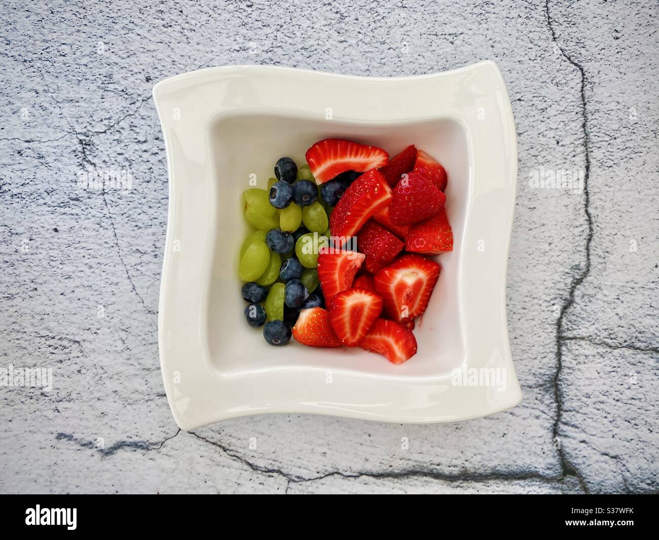 Closeup view of fresh summer berries in a white bowl against a textured natural cracked stone background. Preparing strawberries, blueberries and grapes on the kitchen counter - Smartphone Captured Stock Image