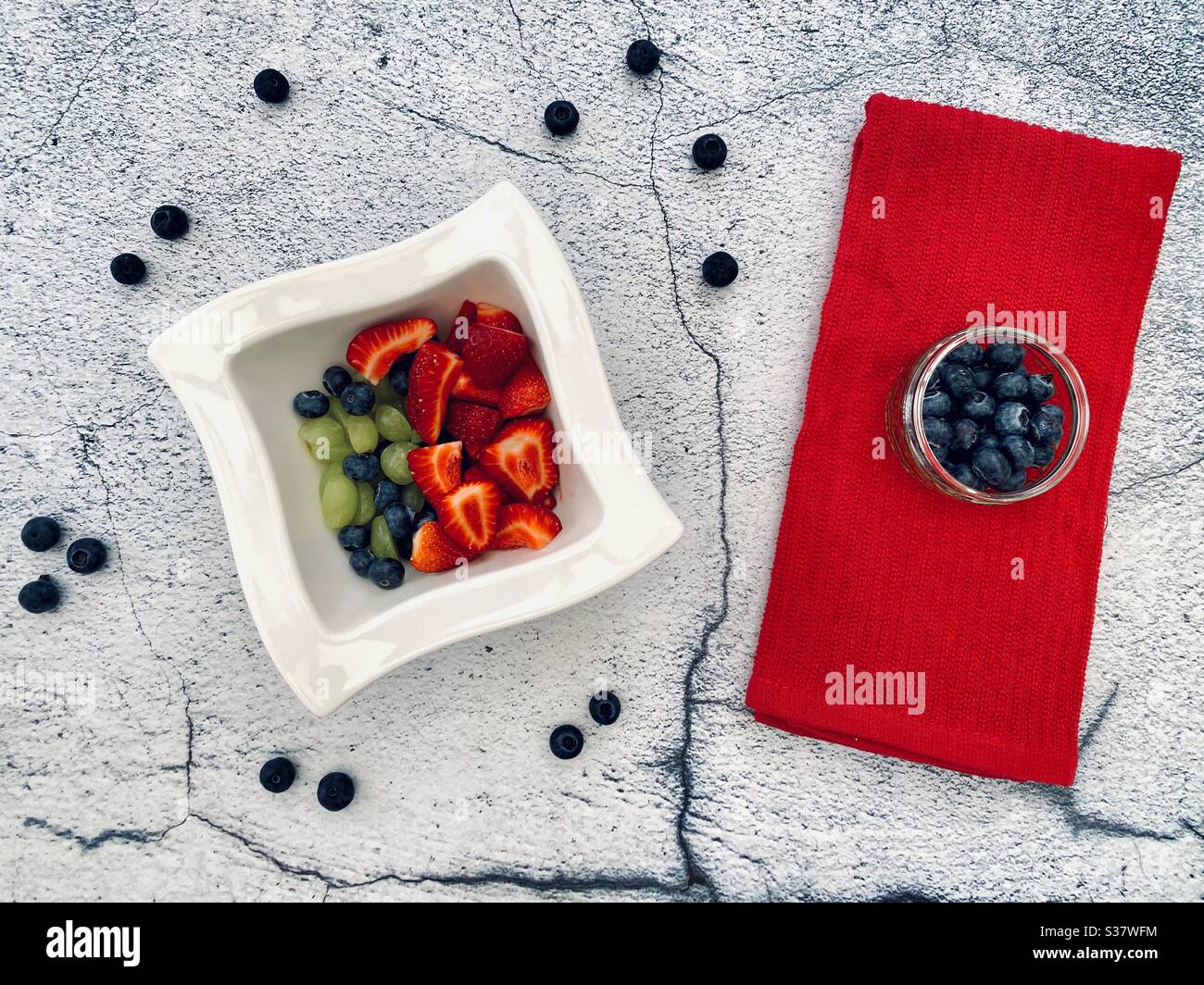 Closeup view of fresh summer berries in a white bowl against a textured natural cracked stone background. Preparing strawberries, blueberries and grapes on the kitchen counter. - Smartphone Captured Stock Image