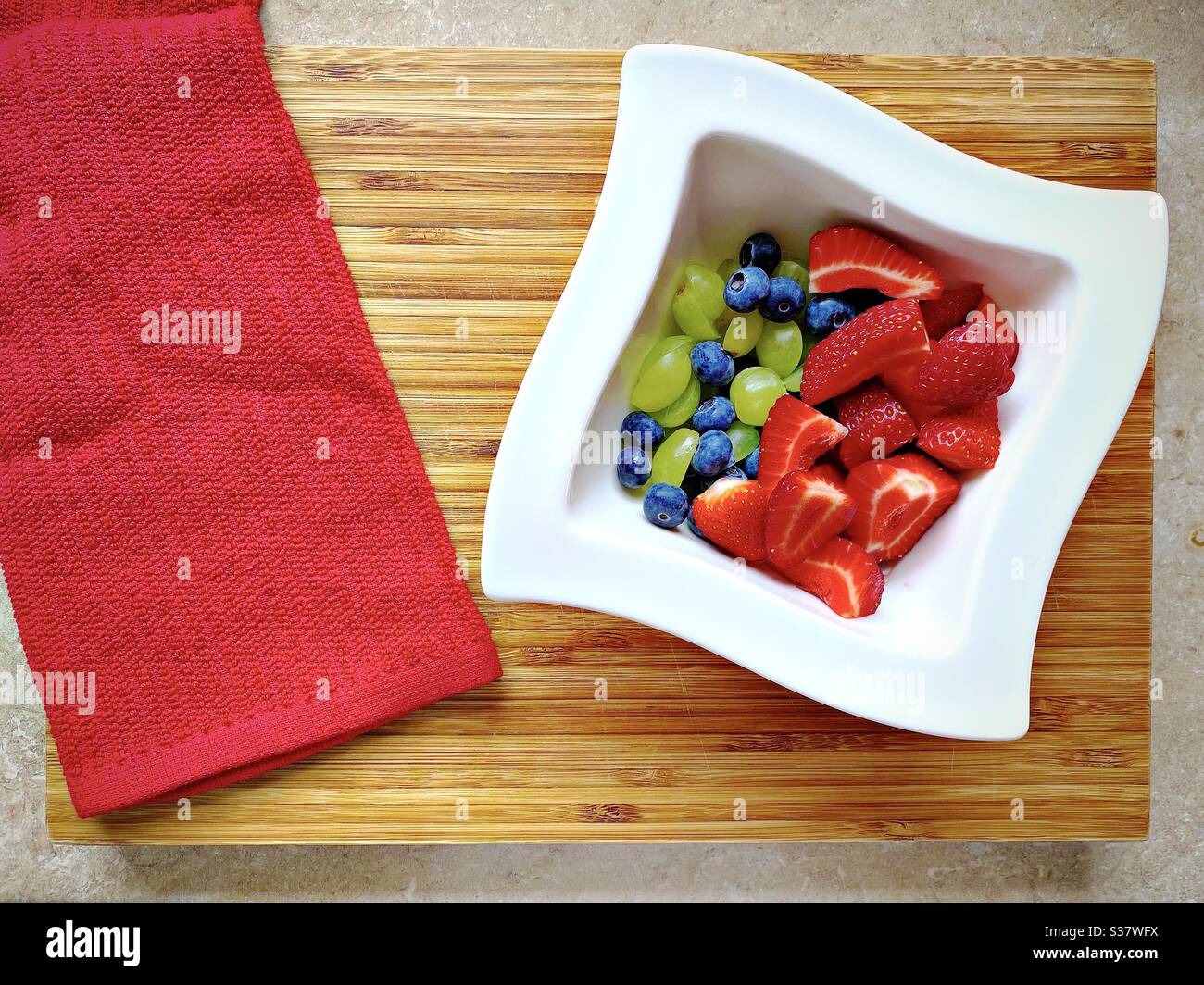Closeup view of fresh summer berries in a white bowl against a rustic wooden chopping board. Preparing strawberries, blueberries and grapes on the kitchen counter. - Smartphone Captured Stock Image