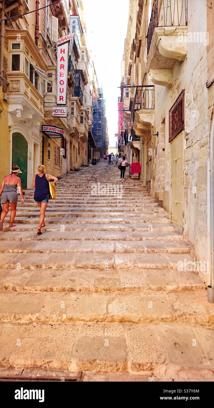 A photograph of a narrow steep street in Valletta, Malta. Tourists and locals browsing the shops and businesses. - Smartphone Captured Stock Image