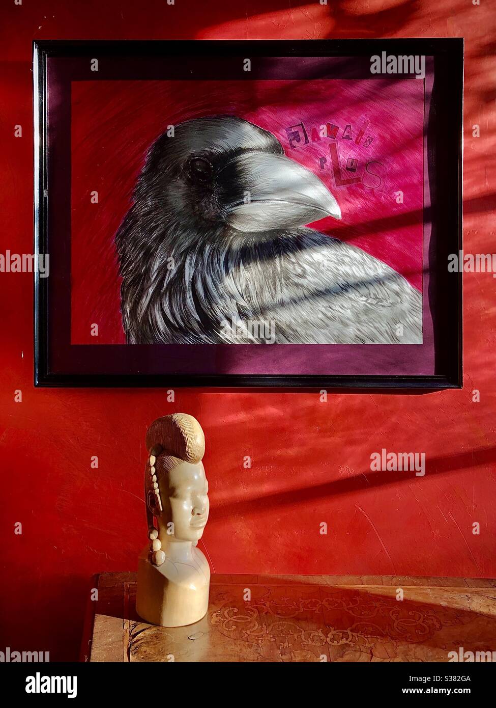 Artist’s drawing of crow and African head sculpture in sunlight against red wall. Stock Photo