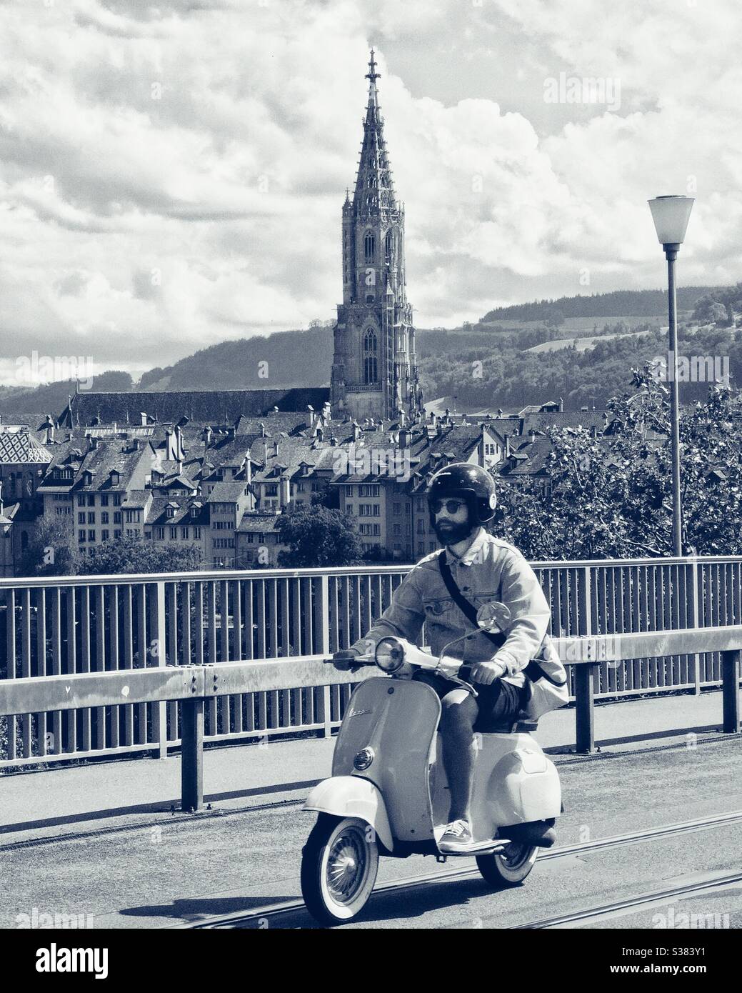 Man on Vespa scooter crosses bridge in Bern, with cathedral tower in background - Smartphone Captured Stock Image