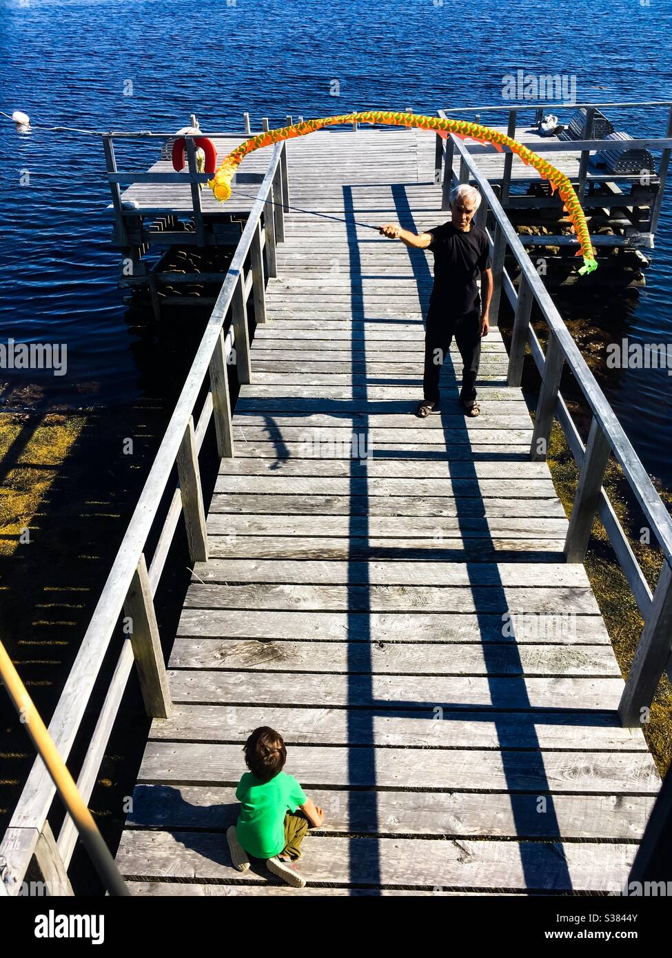 Grandpa flies a kite with toddler watching. Dock overlooking the blue ocean. Sunny day. Nova Scotia, Canada. Simple, classic, low-tech fun, outdoors, open air. - Smartphone Captured Stock Image