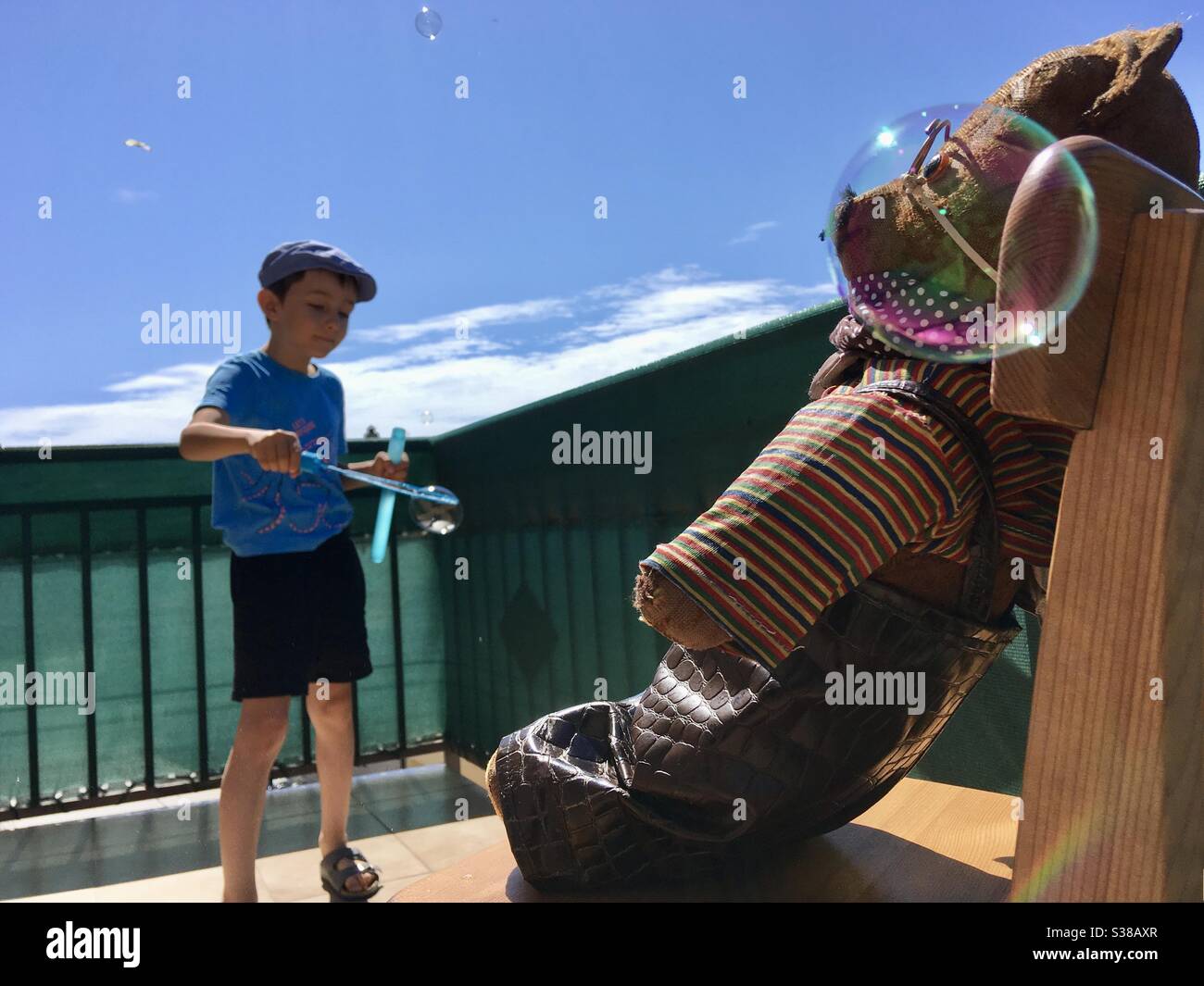 Child kid blowing soap bubbles on balcony with his teddy bear seated on chair - Smartphone Captured Stock Image