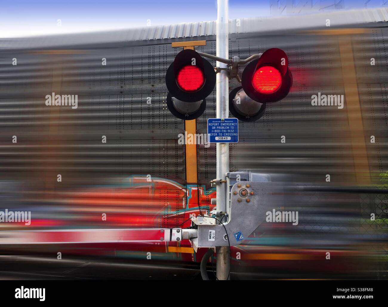 A freight train speeding past a railway crossing, Ontario, Canada. Red lights flashing. Concept, fast and slow, fixed and moving, danger approaching. Risky, caution, transient, fleeting, momentary Stock Photo
