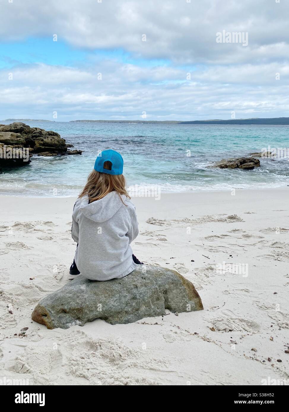 Young girl with long hair, wearing cap and hoodie sits on rock on white sandy beach looking out to sea. Concept of wanderlust, isolation, dreaming and peacefulness. Stock Photo