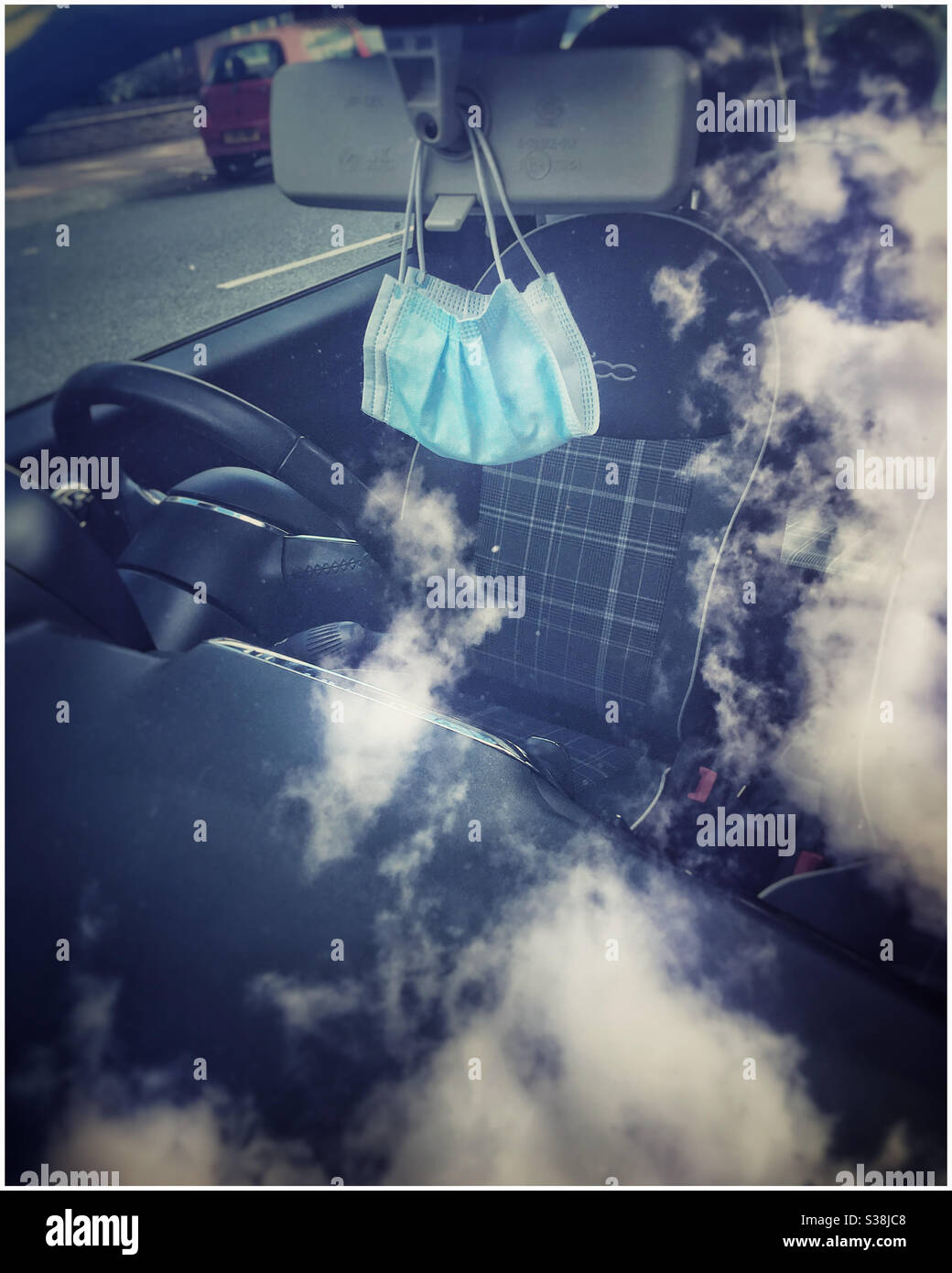 Face mask hangs from rear view mirror, seen through windshield reflecting clouds - Smartphone Captured Stock Image