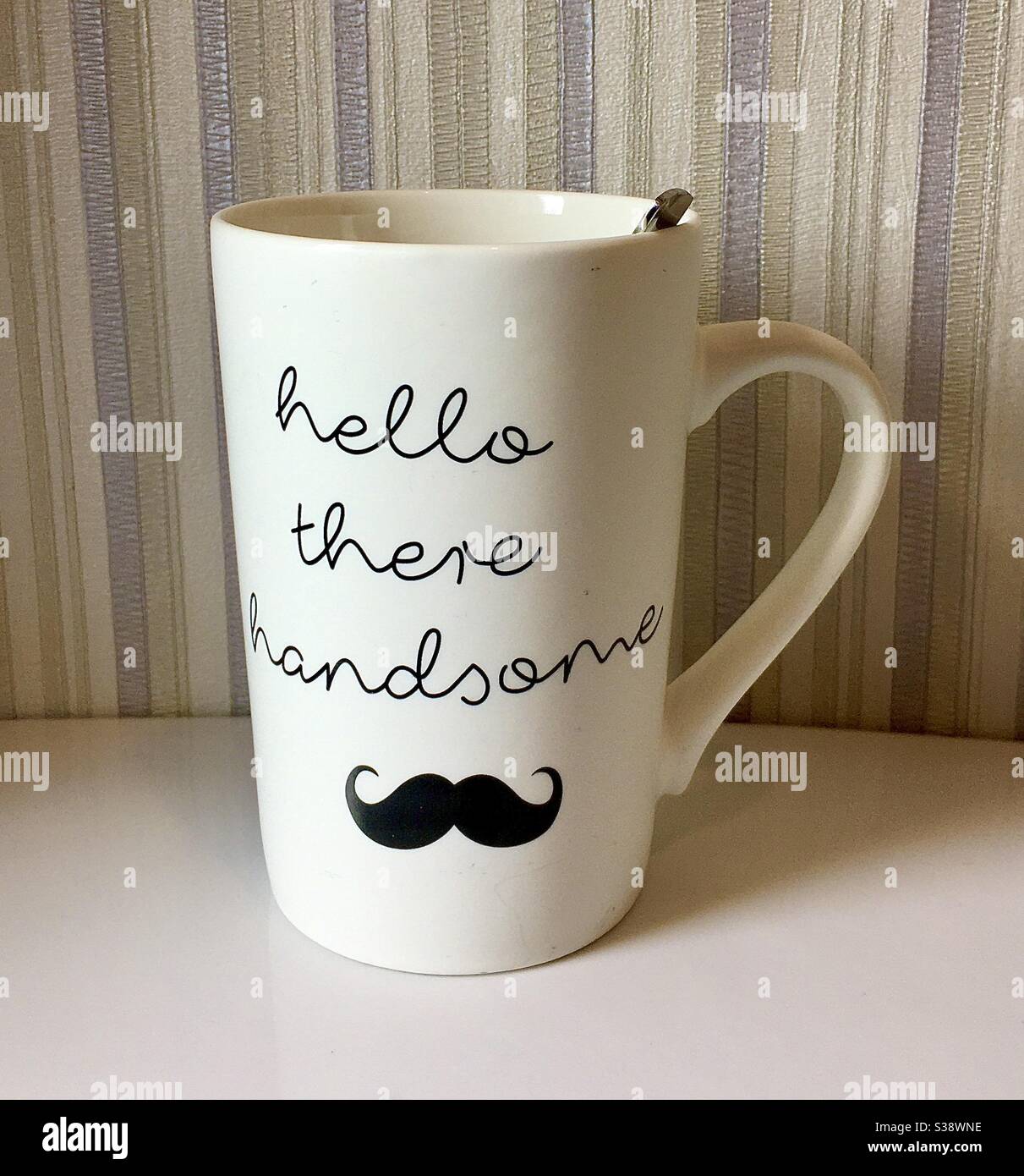 Modern, tall white mug with flattering greeting on the side, together with a picture of a black curly moustache below. Photographed against a vertically-striped wallpaper. Stock Photo