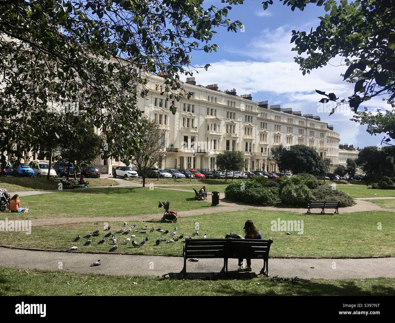 Lady sitting on a bench on a garden square surrounded by Regency architecture in Brighton and Hove, England - Smartphone Captured Stock Image