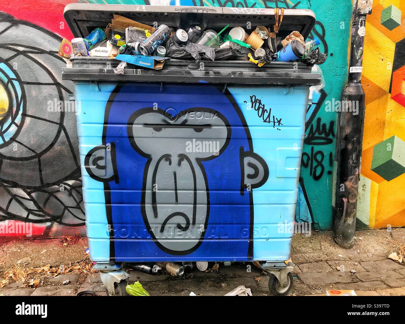 Wheel bin trash can full of garbage or rubbish and painted with graffiti of an angry monkey in street in Brighton UK - Smartphone Captured Stock Image