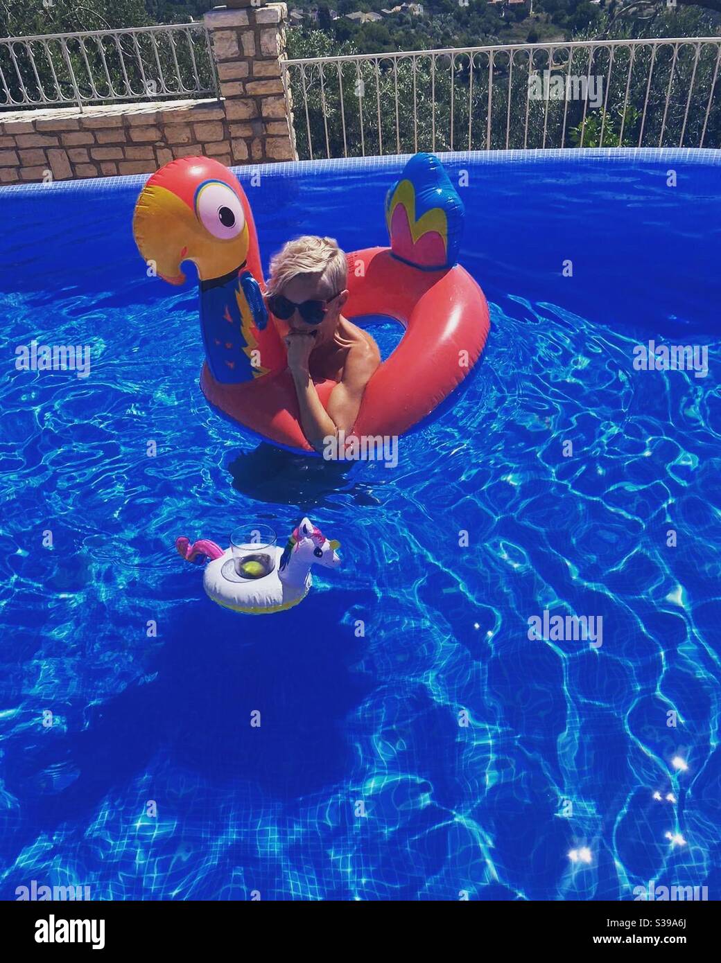 Woman in inflatable parrot biting fist in excitement at inflatable drinks holder in a pool - Smartphone Captured Stock Image