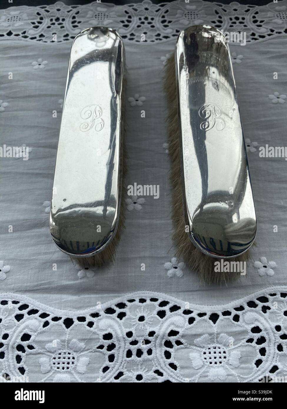A pair of silver hair brushes engraved with the letter B, resting on a broiderie anglais cloth. Stock Photo