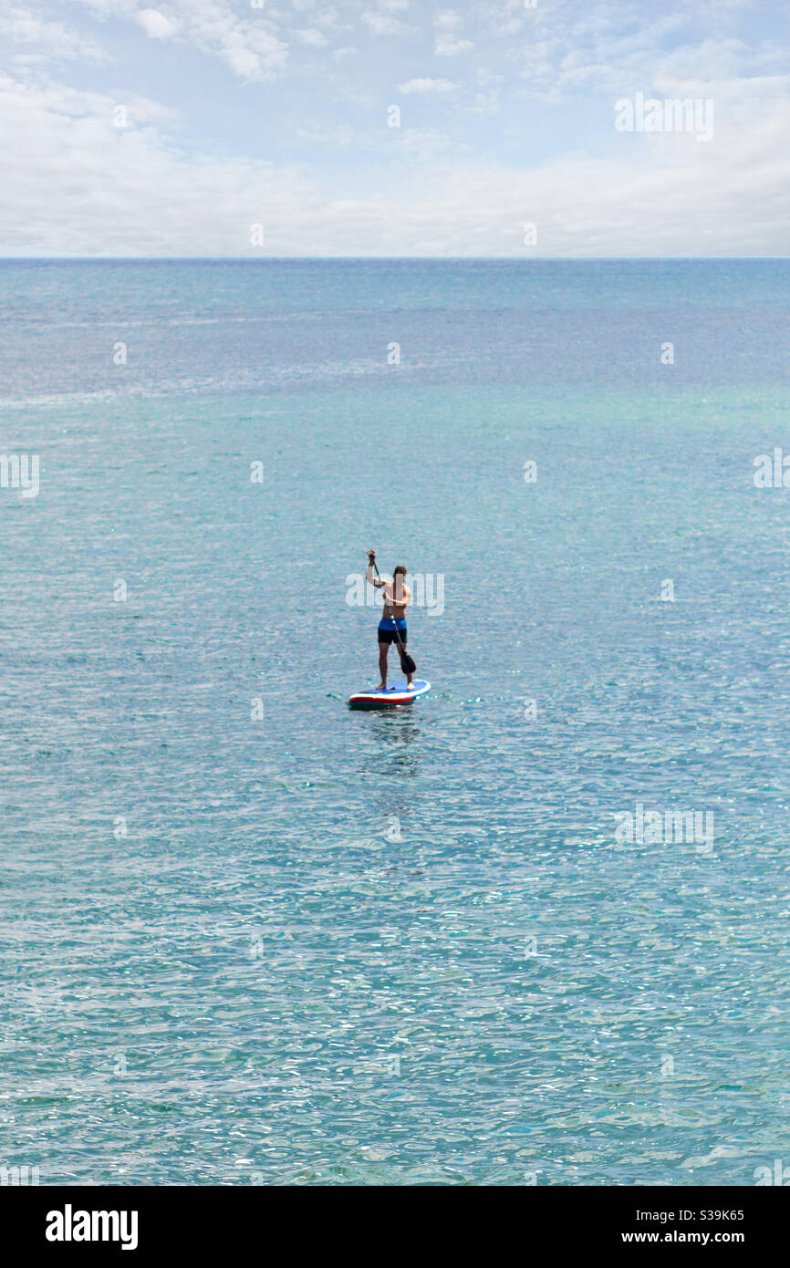 a stand up paddler SUP with blue swimming trunks on the open sea on the blue turquoise water and under a slightly cloudy sky Stock Photo