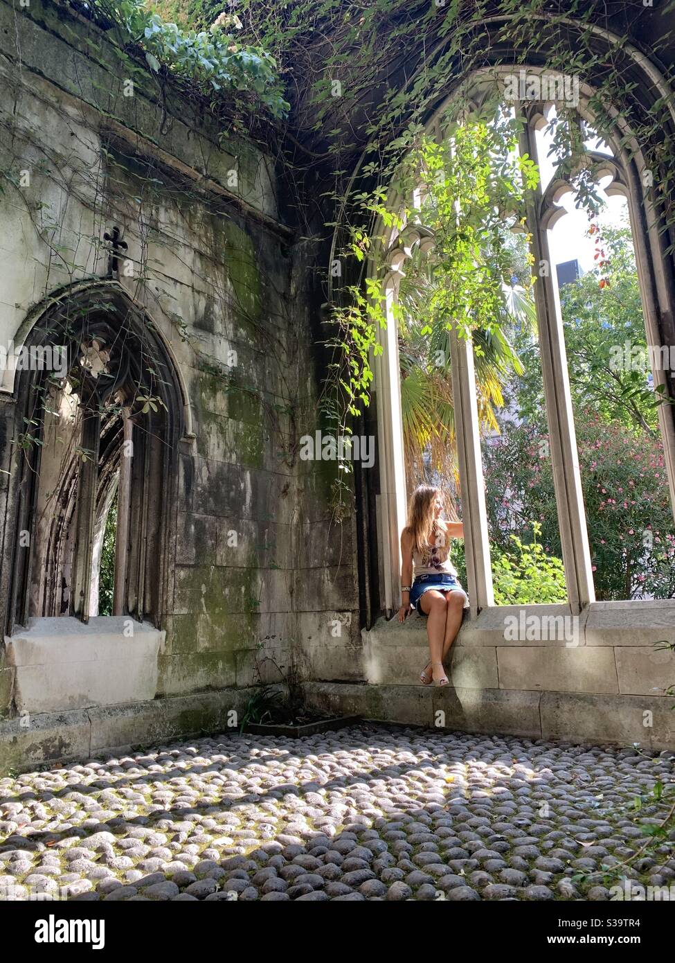Woman sitting in stone archway with plants and sun shining through - Smartphone Captured Stock Image