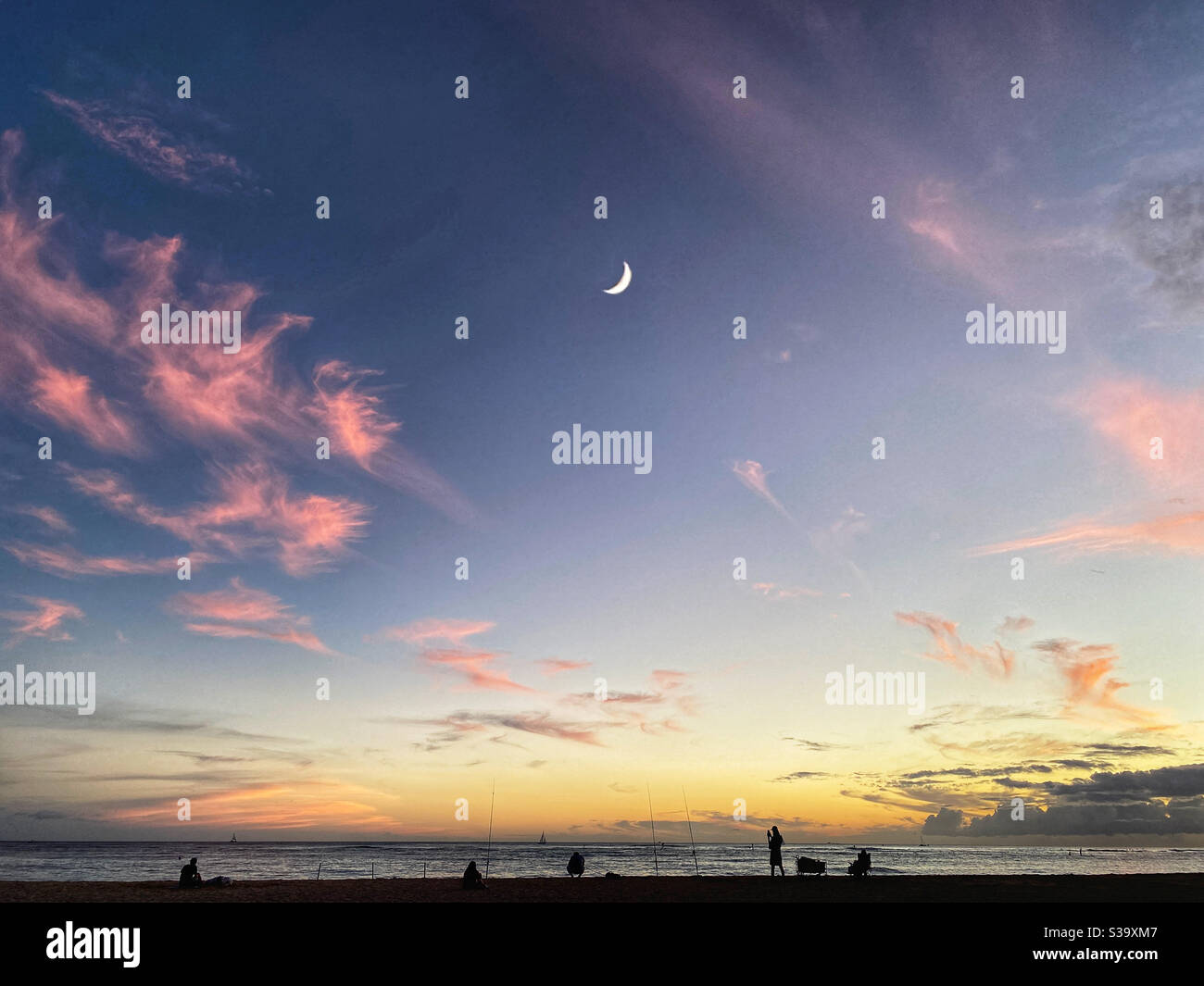 A crescent moon over silhouetted figures fishing on a beach at sunset Stock Photo