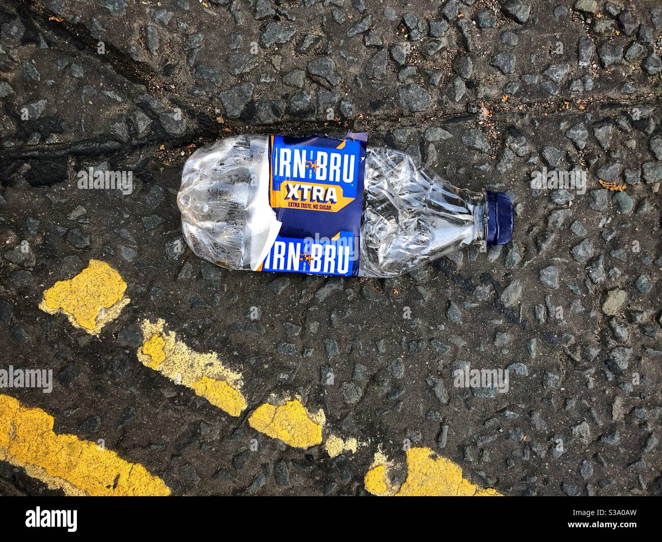 A plastic Irn Bru Extra bottle lies discarded on the road tarmac in Dumfries, Scotland, damaged yellow line road markings visible. - Smartphone Captured Stock Image