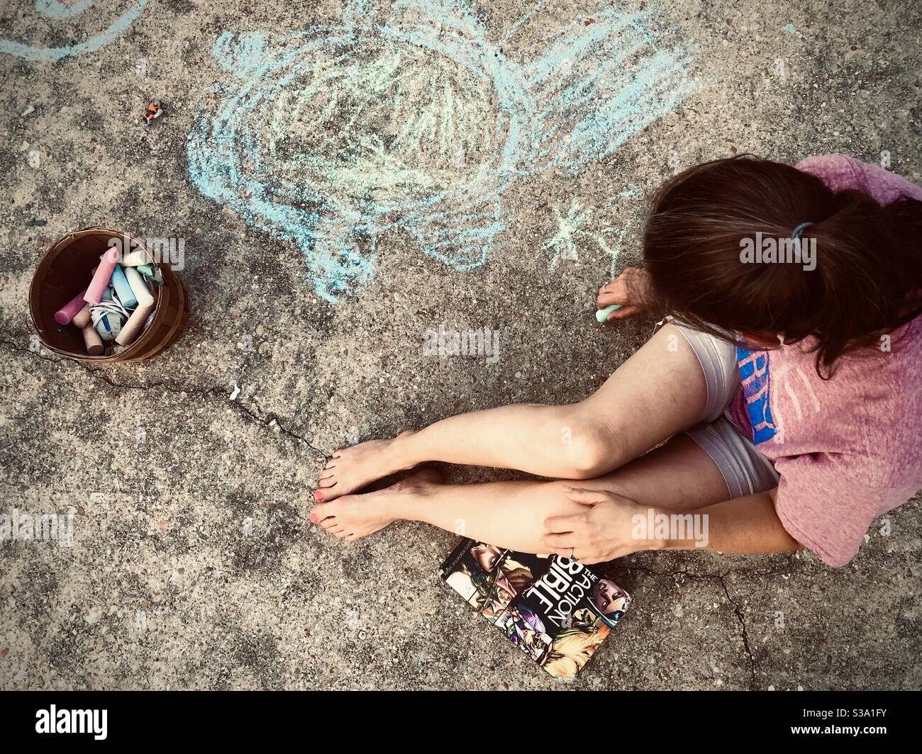 Using chalk to draw on her driveway is Holly Tippett. Photo take July 19, 2020 in Columbus, Mississippi. A book labeled “The Action Bible” rests near the bottom leg. - Smartphone Captured Stock Image