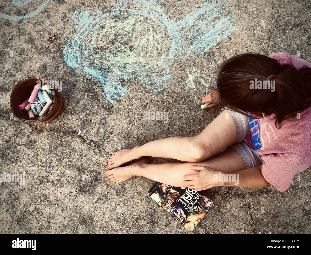 Using chalk to draw on her driveway is Holly Tippett. Photo take July 19, 2020 in Columbus, Mississippi. A book labeled “The Action Bible” rests near the bottom leg. Stock Photo