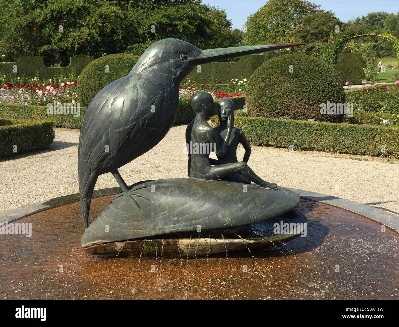 Fountain sculpture of oversized bird with 2 figures sitting on a mussel shell with topiary in the background Stock Photo