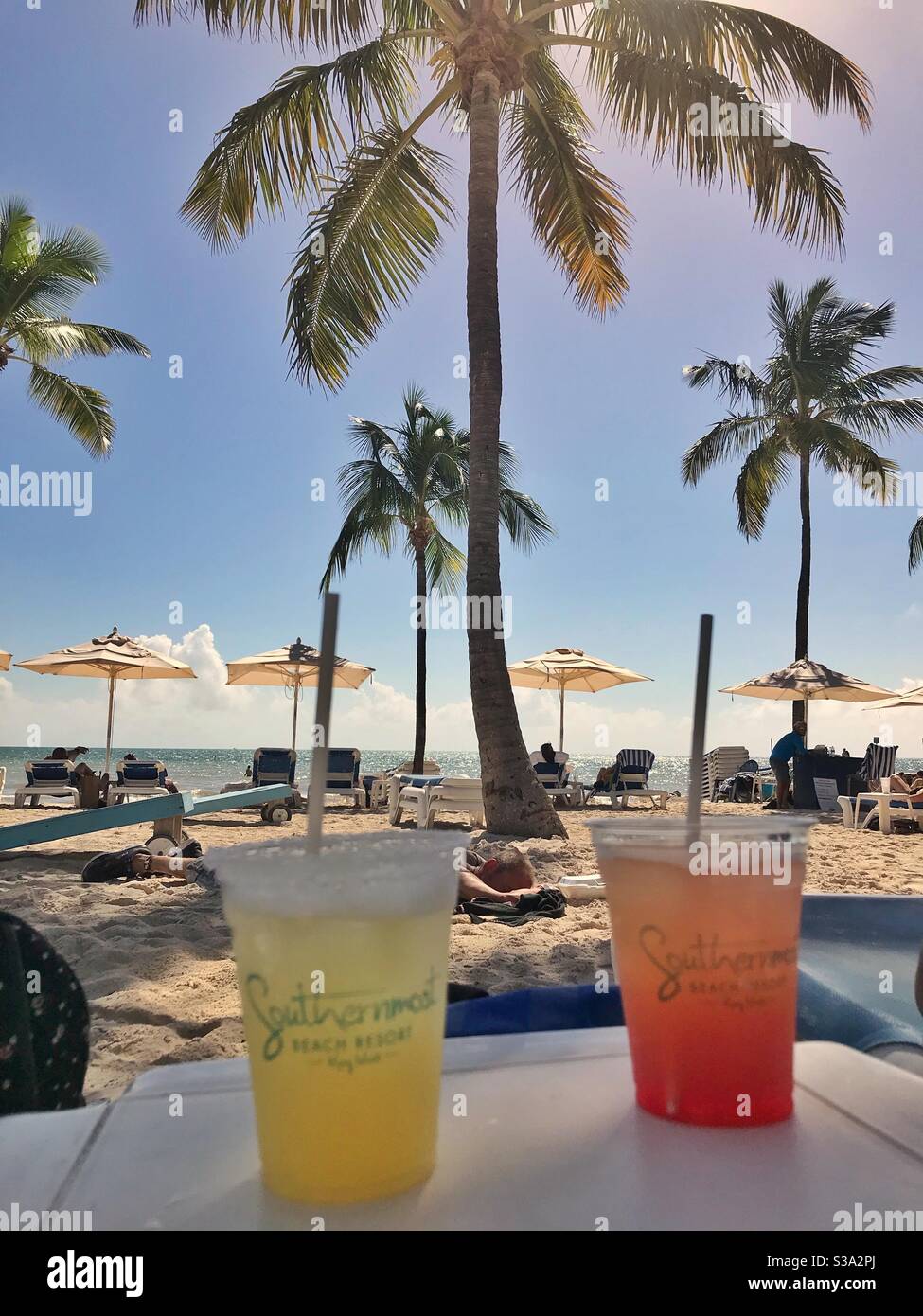 Two cold drinks on a table by the beach with palm trees and umbrellas