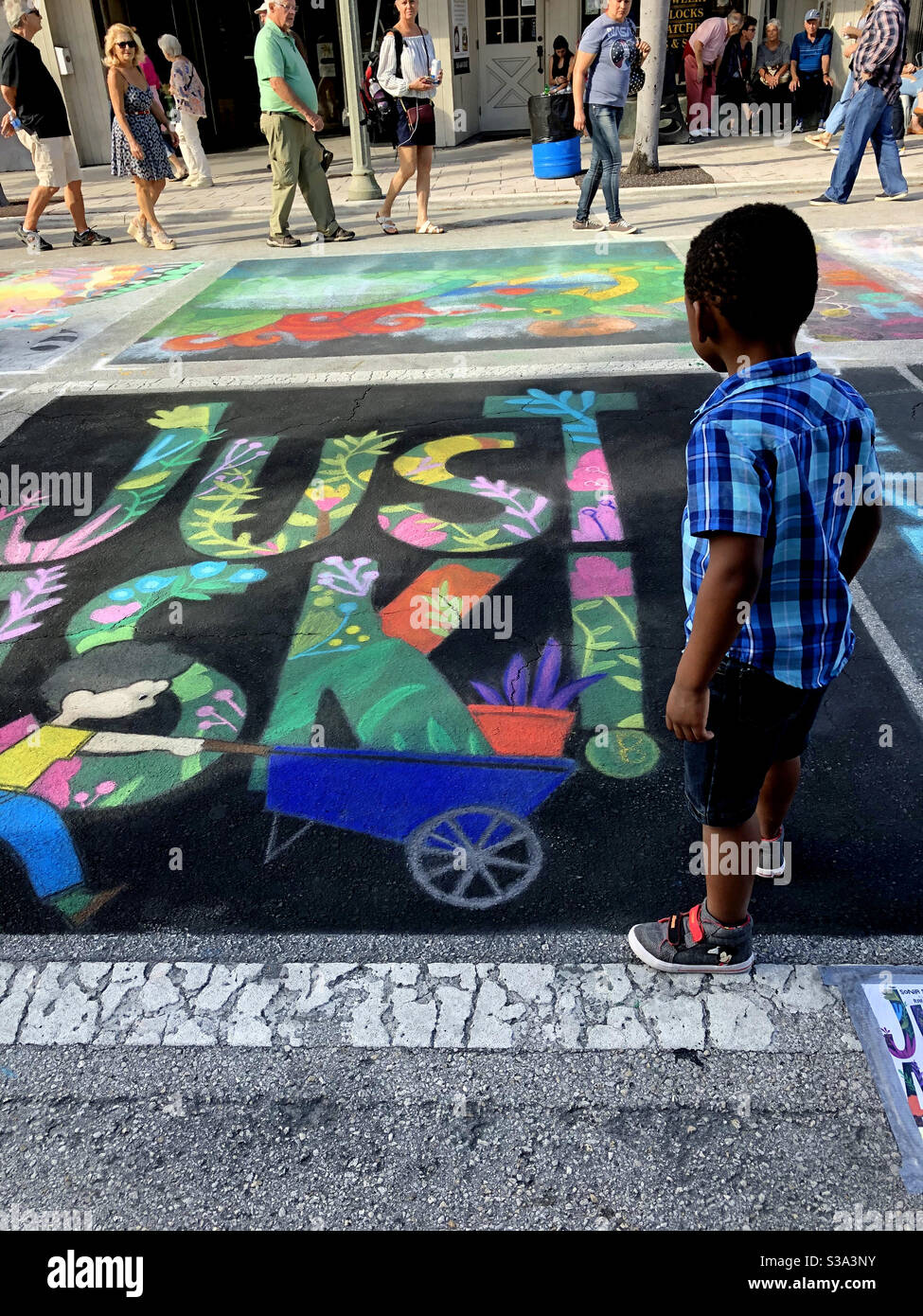 Little boy reading a colorful chalk sign “Just Ask!” at a community