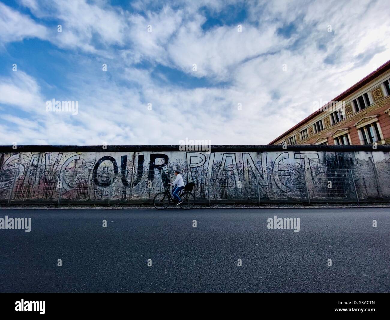A woman rides her bike along a part of the Berlin Wall that is still remaining up. Graffiti on the wall reads “save our planet.”  Berlin, Germany. Stock Photo