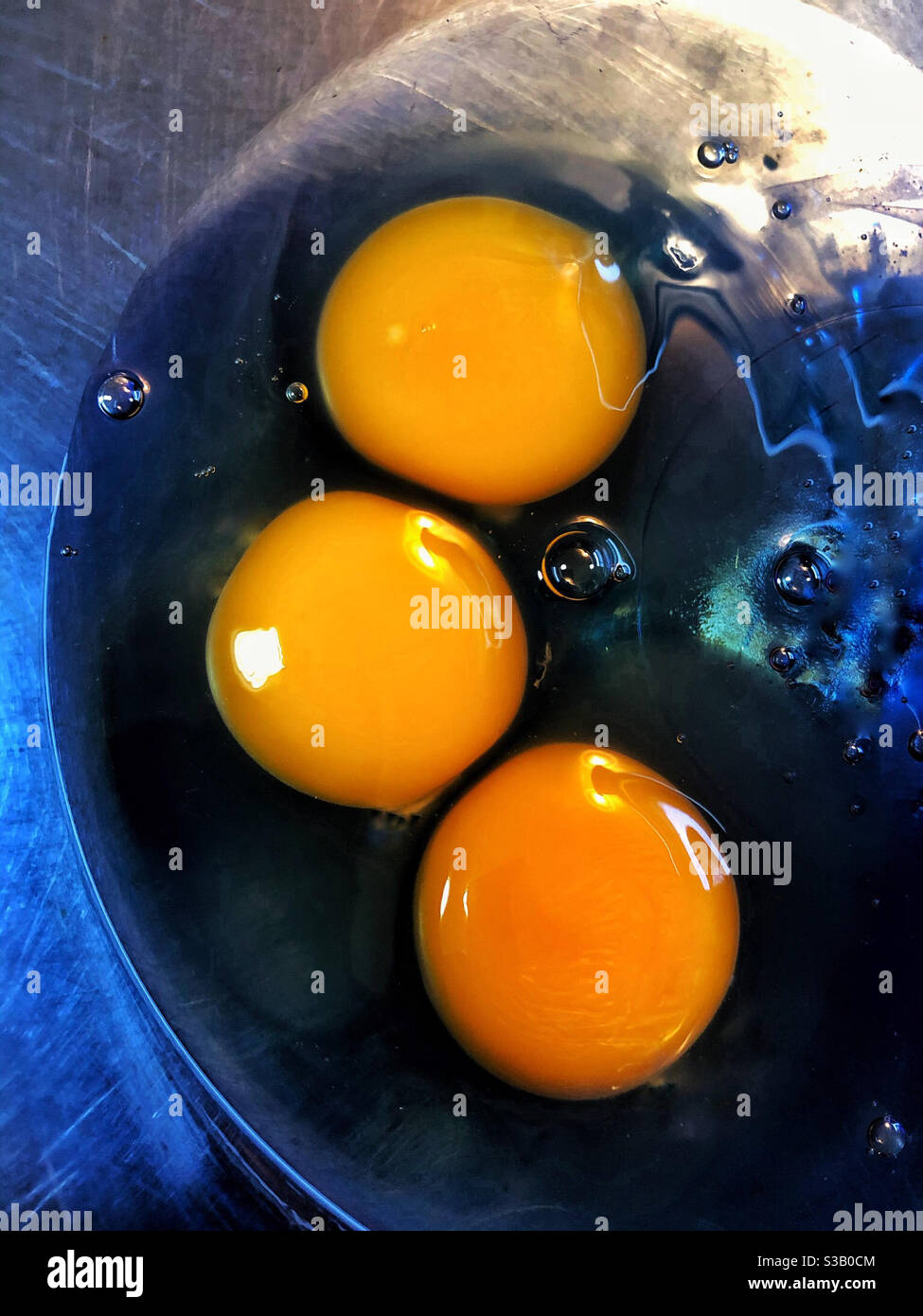 Three raw egg yolks in blue glass mixing bowl, surrounded by the clear, uncooked egg whites on stainless steel table - Smartphone Captured Stock Image