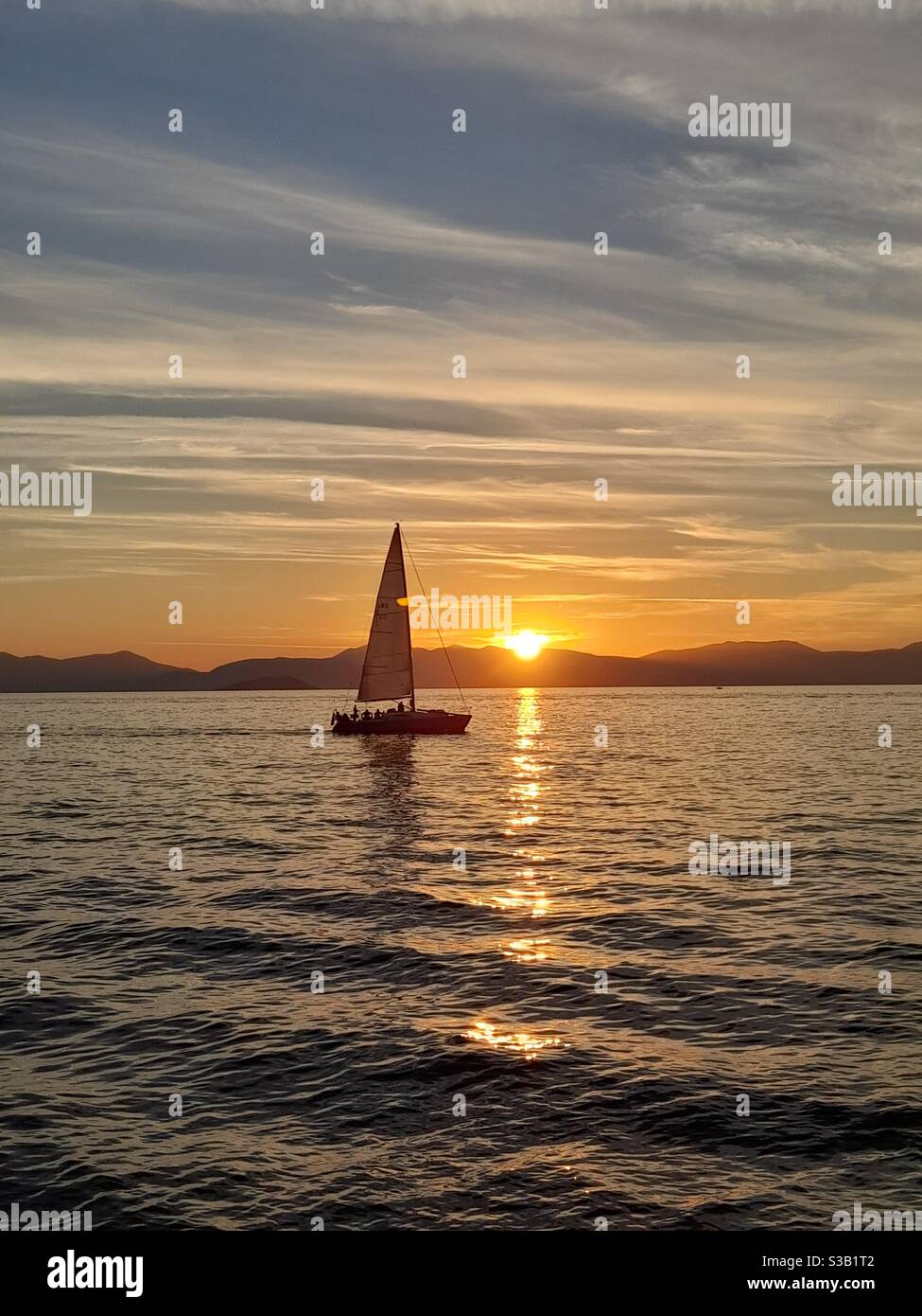 Sailboat gliding over the calm sea at sunset, golden reflections on the water with a dramatic sky, creating a serene and idyllic seascape. - Smartphone Captured Stock Image