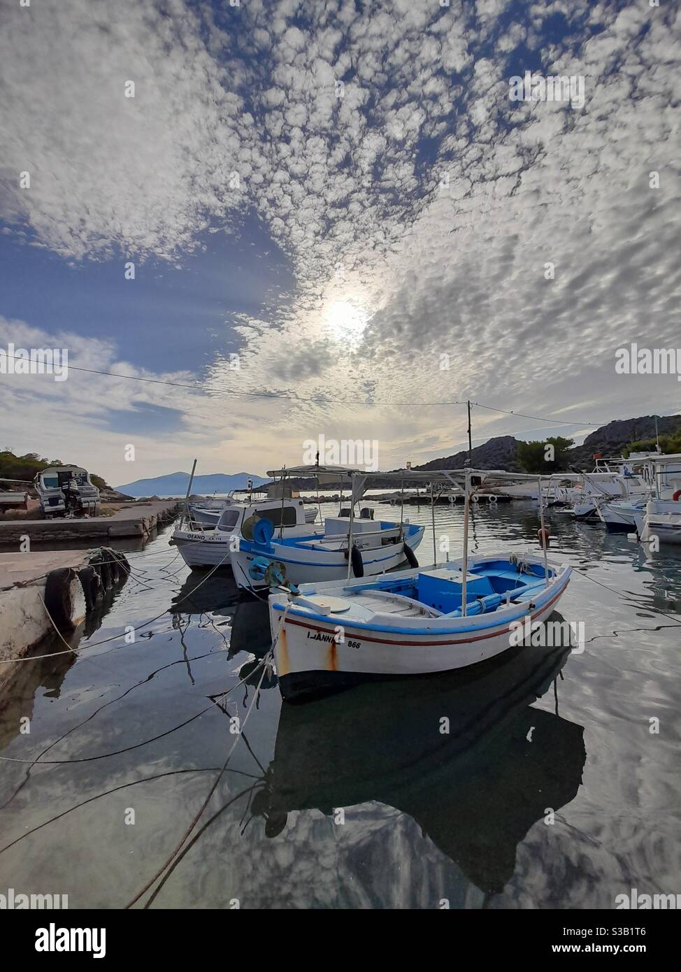 Small fishing boats gently float on still waters, reflecting a textured sky and sunlight in a peaceful harbor setting, in Agkistri island, Greece - Smartphone Captured Stock Image