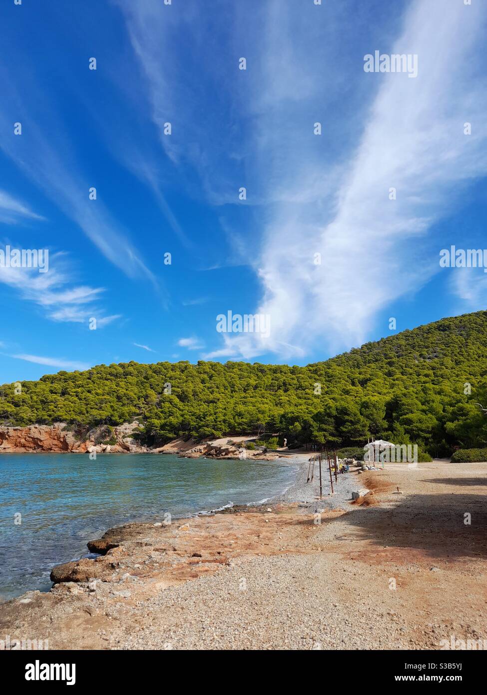 A serene, secluded beach on Agkistri Island, Greece, surrounded by lush green hills and crystal-clear waters under a vibrant blue sky with wispy cloud - Smartphone Captured Stock Image