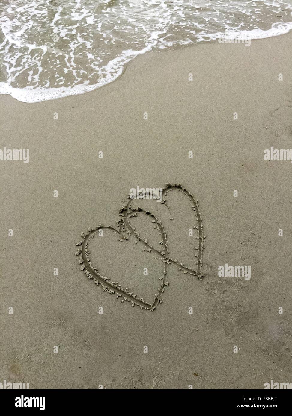Two hearts drawn in light black sand on the beach, a wave coming. Symbol of new love, renewal, turbulence ahead, ephemera, or a transient interaction, Nova Scotia, Canada - Smartphone Captured Stock Image