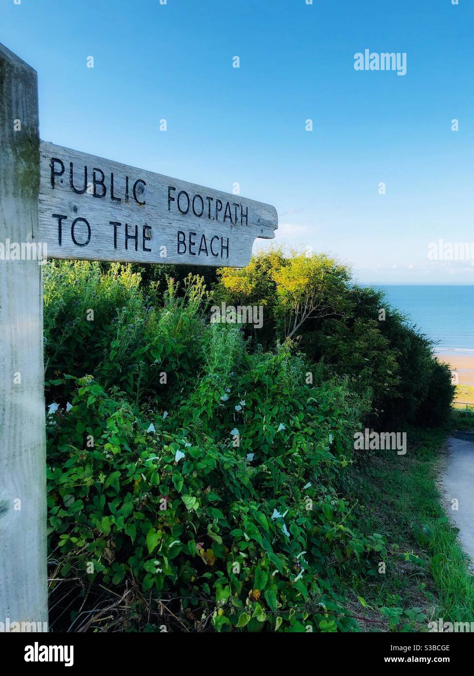 Wooden sign pointing to a public footpath to the beach with a glimpse of the sea in the background - Smartphone Captured Stock Image
