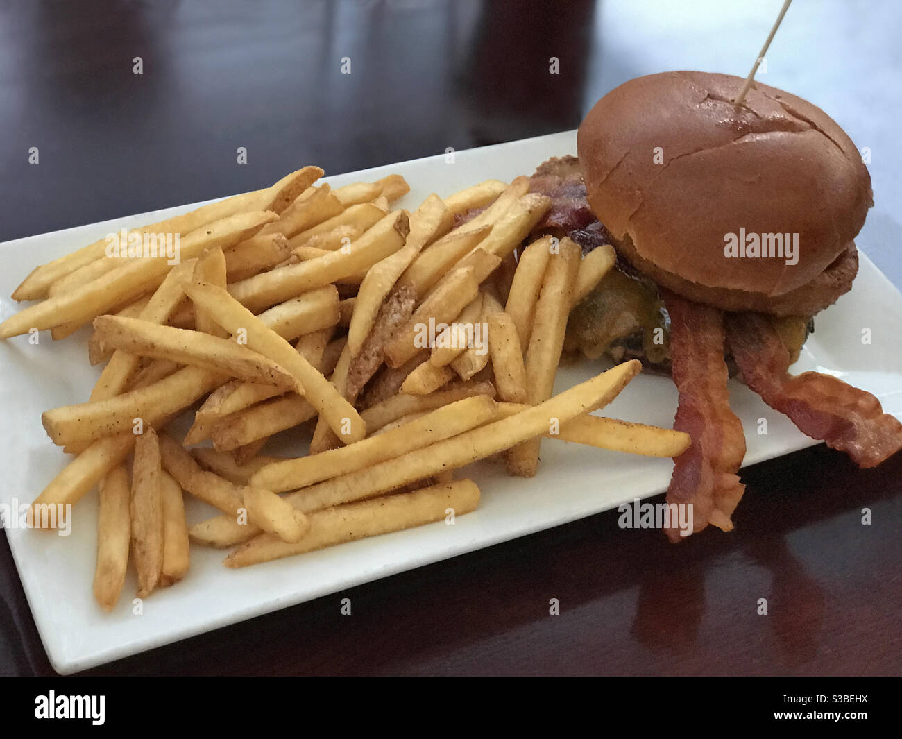 A gourmet BBQ cheeseburger, topped with bacon and onion rings, is shown with French fires plated on a restaurant table. - Smartphone Captured Stock Image