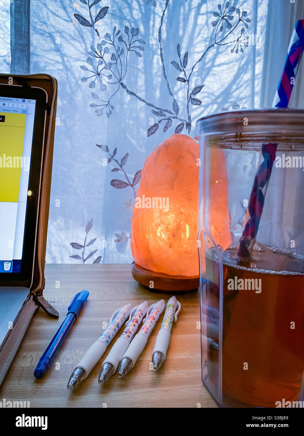 A writer’s desk with laptop, an assortment of pens, a travel glass of ice tea with a reusable straw and a Himalayan salt lamp. - Smartphone Captured Stock Image