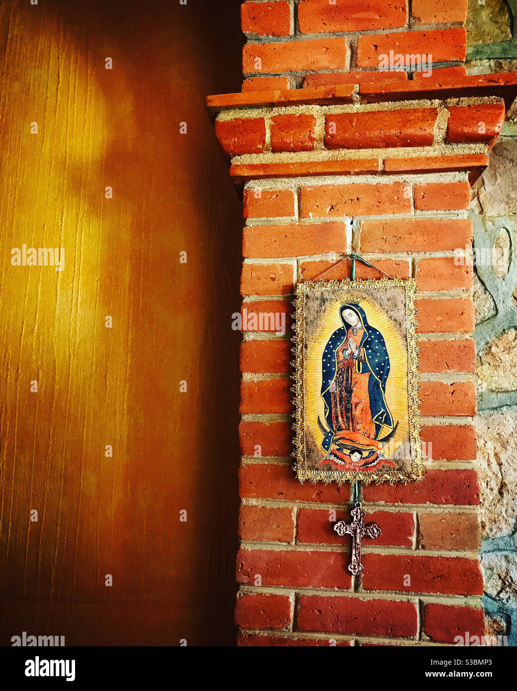 A banner of Our Lady of Guadalupe and a cross decorate a red bricks column in a house with orange walls in a home in Peña de Bernal, Queretaro, Mexico - Smartphone Captured Stock Image