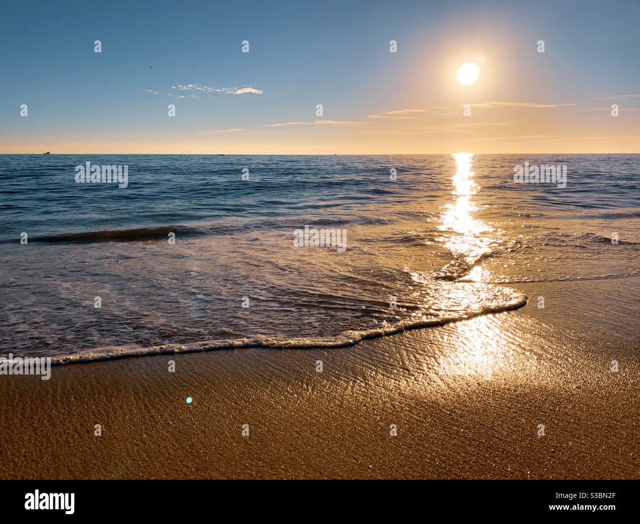 Low angle shot of a gentle wave washing onto sandy beach with late afternoon sun low on the horizon illuminating the ocean. Stock Photo