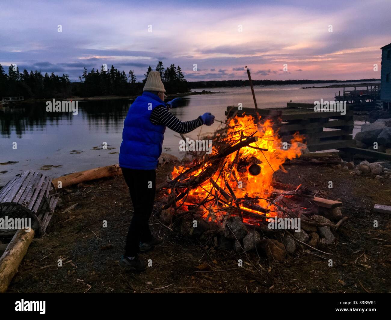 A woman tends to a bonfire by the sea at sunset on a chilly evening. A quick, safe method for disposal of scrap wood outdoors. - Smartphone Captured Stock Image