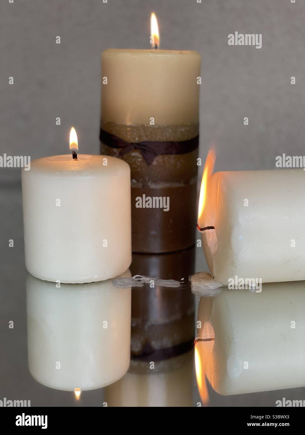 Three burning candles one turned sideways with wax dripping on mirrored surface Stock Photo