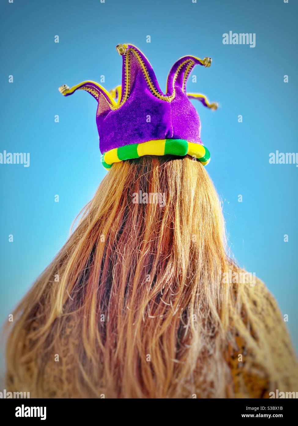 Blonde Woman photographed from behind wearing a felt Mardi Gras crown . Shrove Tuesday or Fat Tuesday  a party before Ash Wednesday  blue sky back ground Stock Photo