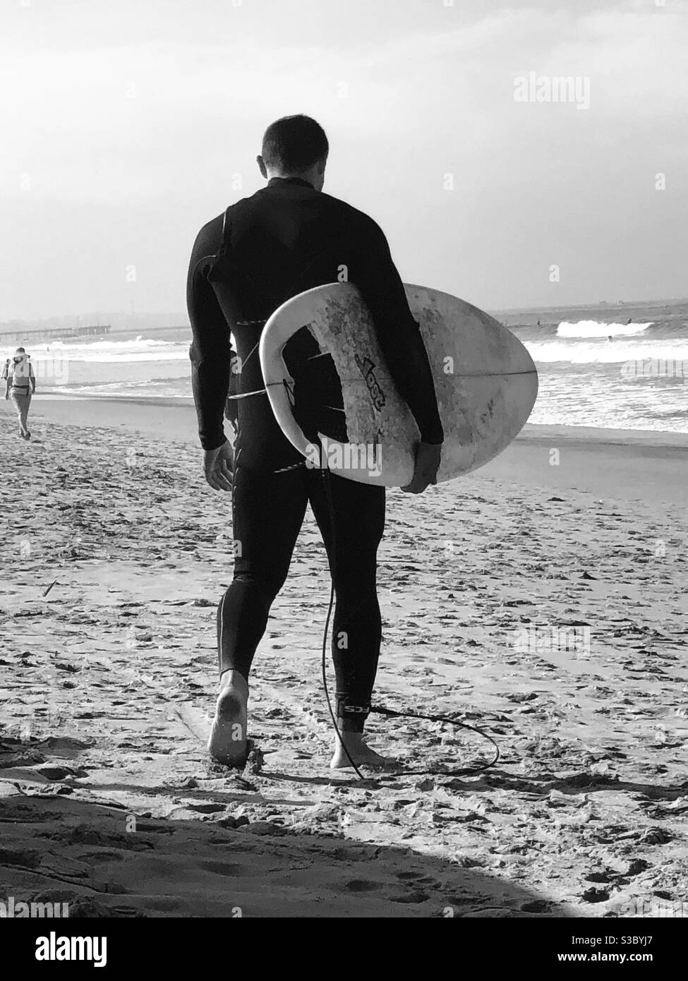 Black and white photo of a surfer caring a surfboard on the beach in Santa Monica headed toward the waves of the Pacific ocean. Stock Photo