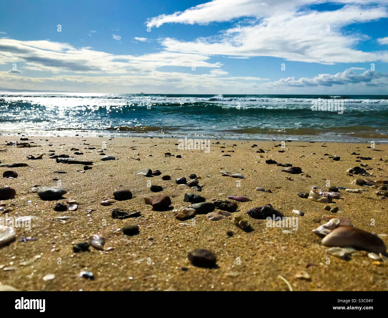 Close view of the beach that leads to the sea in the background. The beach is full of small shells and pebbles that the ocean placed on the beach. There is a cloudy sky and blue waters Stock Photo