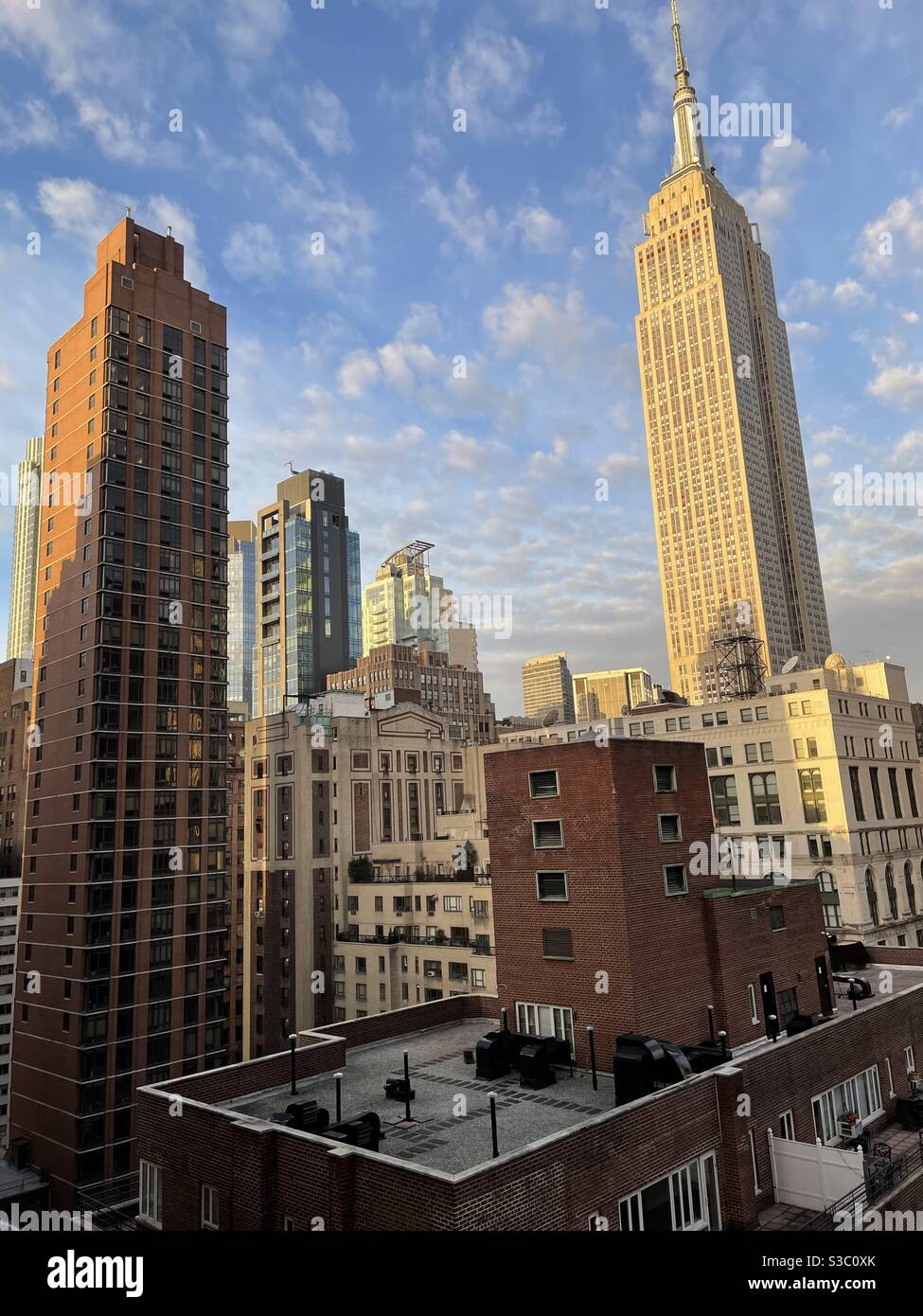 Dramatic view of the Empire State building and other skyscrapers and high-rise buildings in Midtown Manhattan, including a rooftop & fluffy clouds in a blue sky. - Smartphone Captured Stock Image