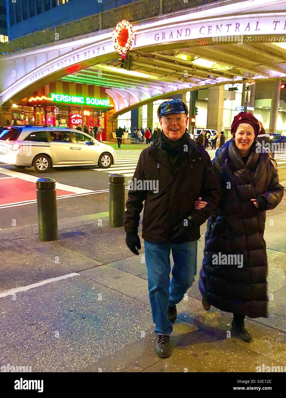 Dressed in warm clothing & hats on a cold winter night, a man & woman stroll cheerfully arm in arm by Grand Central on 42nd Street in New York City, USA. - Smartphone Captured Stock Image