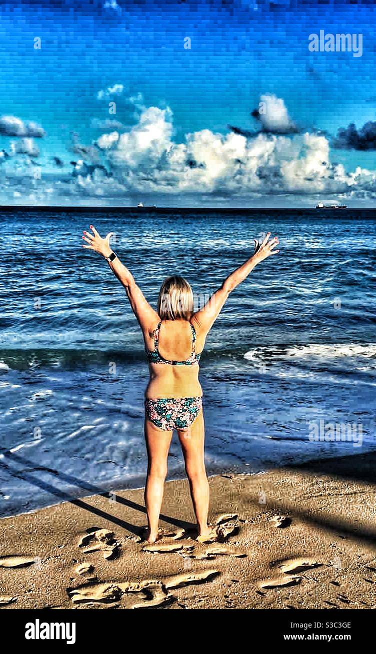 Woman standing on a beach looking out to the Caribbean sea with her arms stretched up to the sky - Smartphone Captured Stock Image