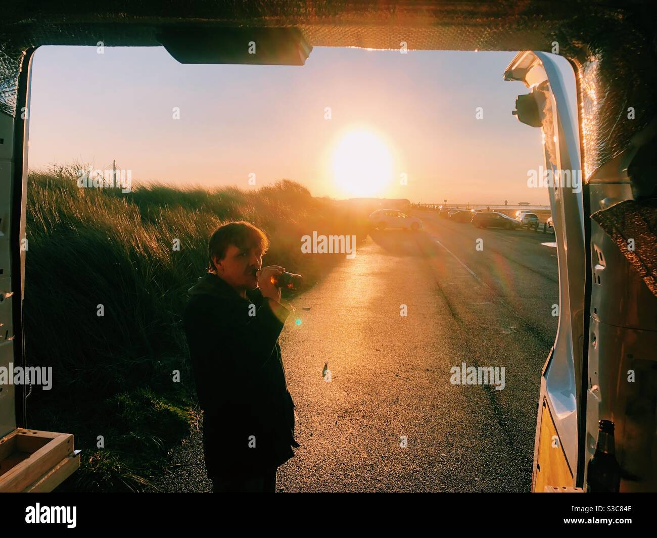 Man drinking beer in front of sunset viewed from back of a camper van - Smartphone Captured Stock Image