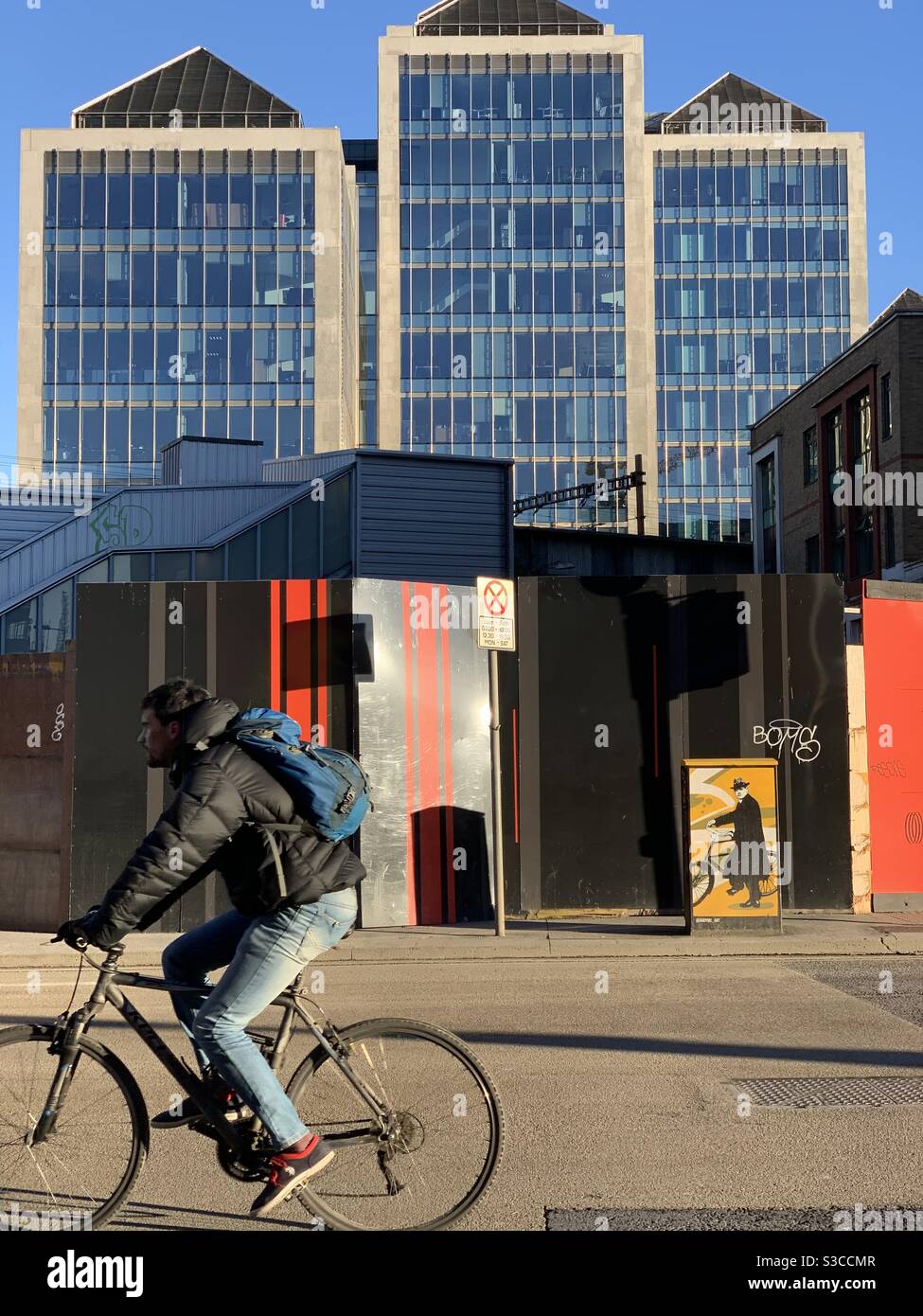 A man on a bicycle passes by a building site hoarding in Dublin. In the background is UlsterBank’s iconic George’s Plaza office complex. Near the hoarding is a mural of Michael Collins with a bicycle. - Smartphone Captured Stock Image