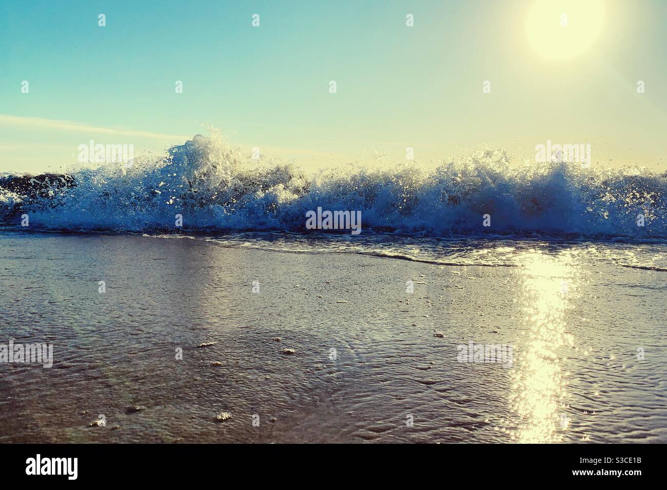 Low angle shot of waves rolling onto sandy beach and splashing water on a sunny afternoon with sunlight reflecting on the wet sand. - Smartphone Captured Stock Image